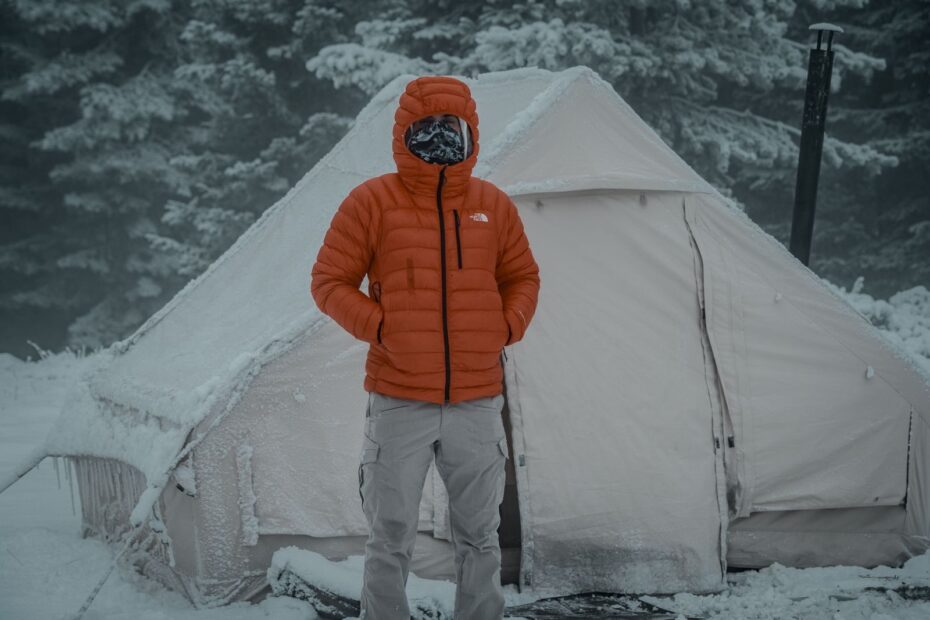 Man in winter jacket standing by a snow-covered tent in a serene outdoor setting.